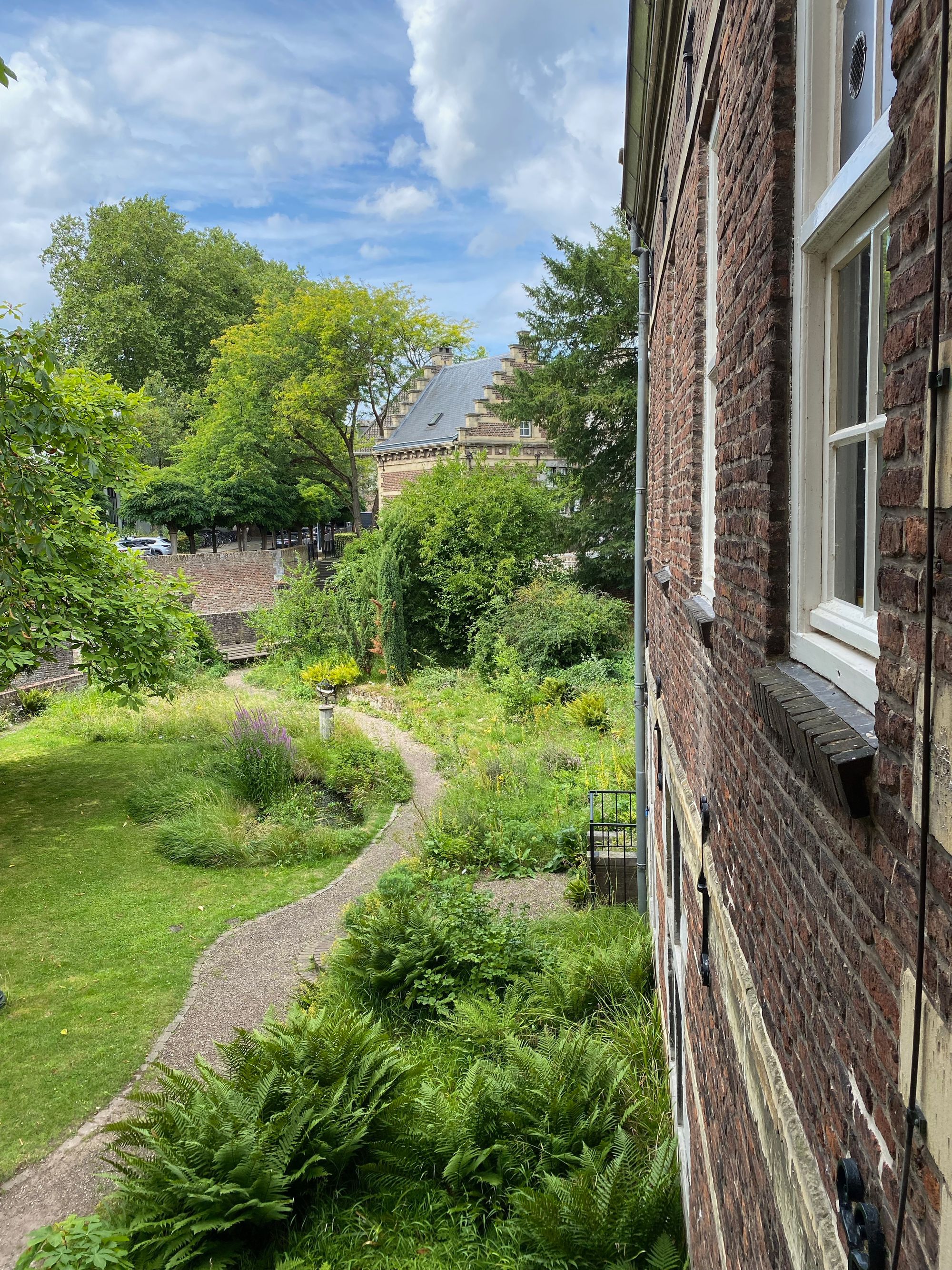 Das Bild zeigt einen Garten mit Farn im Vordergrund und einem Kiesweg, der sich durch das Grün der Wiese schlängelt. Rechts im Bild ist das Gemäuder des Gebäudes zu sehen. 