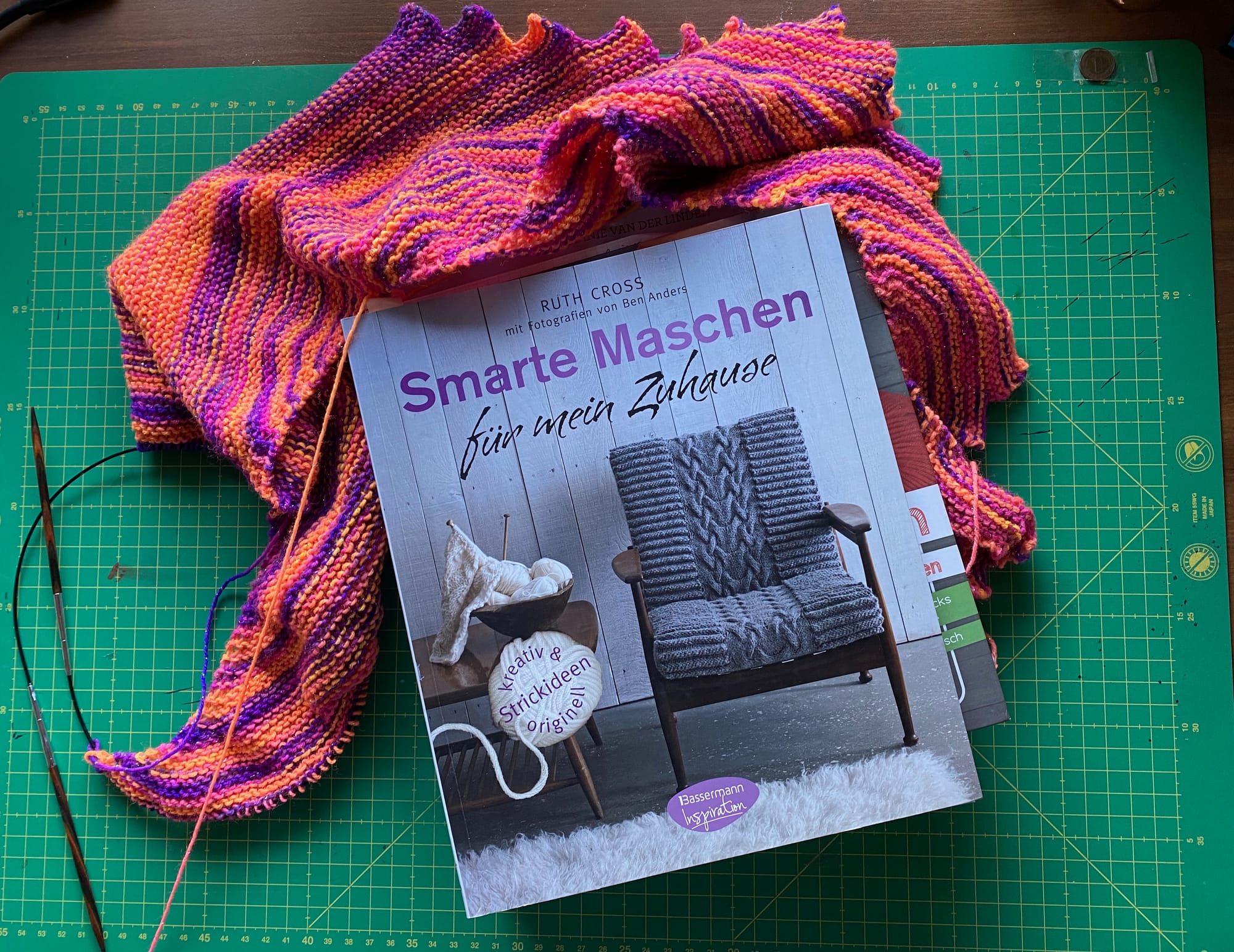 Ein Buch von oben fotografiert. Das Buch liegt auf einer Schneidematte und als Deko wurde ein gestrickter Schal um das Buch drapiert. Der Titel des Buches ist "Smarte Maschen für mein Zuhause"