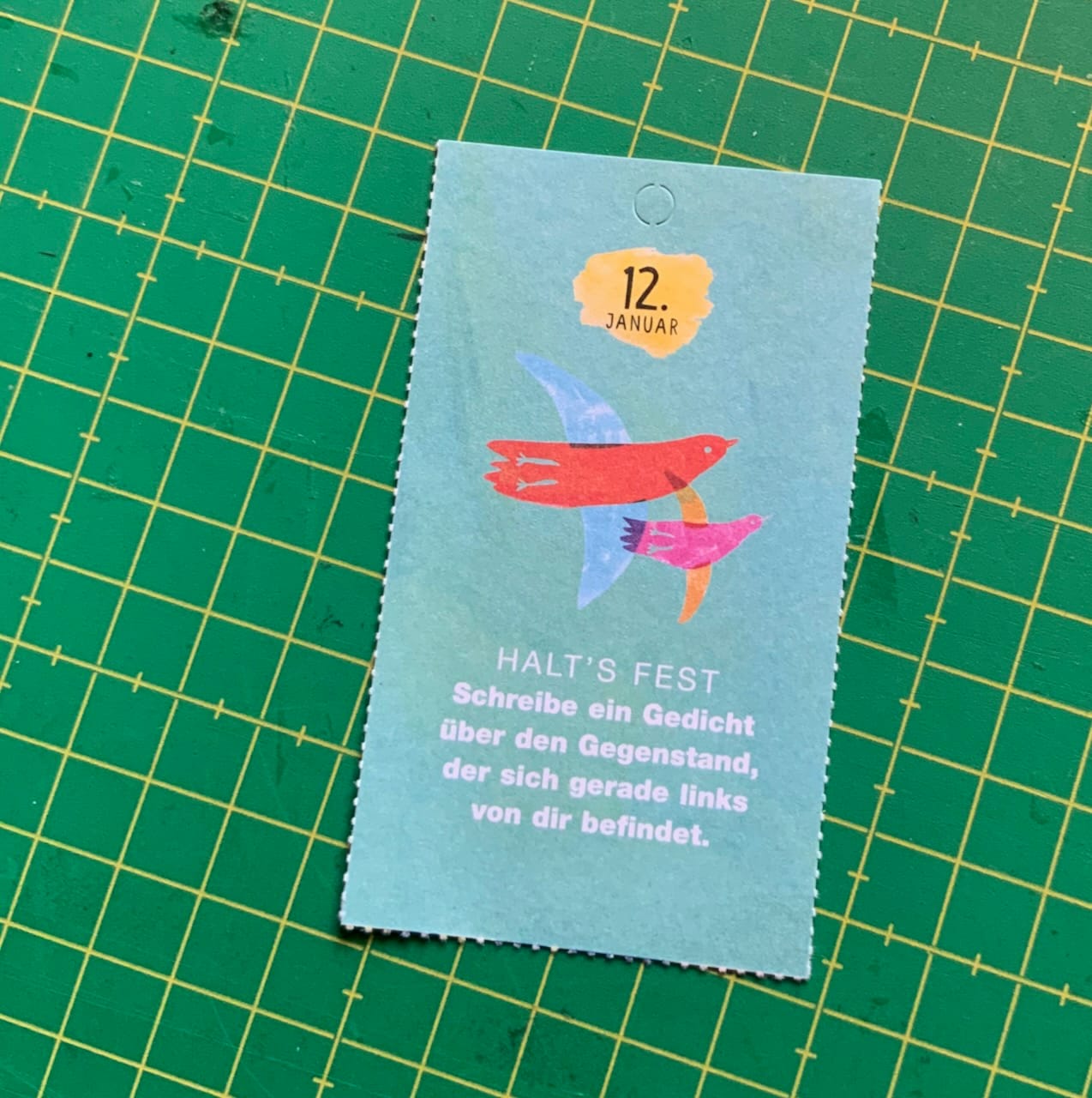 eine rechteckige kleine Karte mit Performations-Markierungen ringsum liegt auf einer grünen Schneidematte. Auf der Karte steht: "12. Januar - Halt's fest - Schreibe ein Gedicht über den Gegenstand, der sich gerade links von dir befindet." Zwischen dem Datum und dem Text sind zwei illustrierte, bunte Vögel.