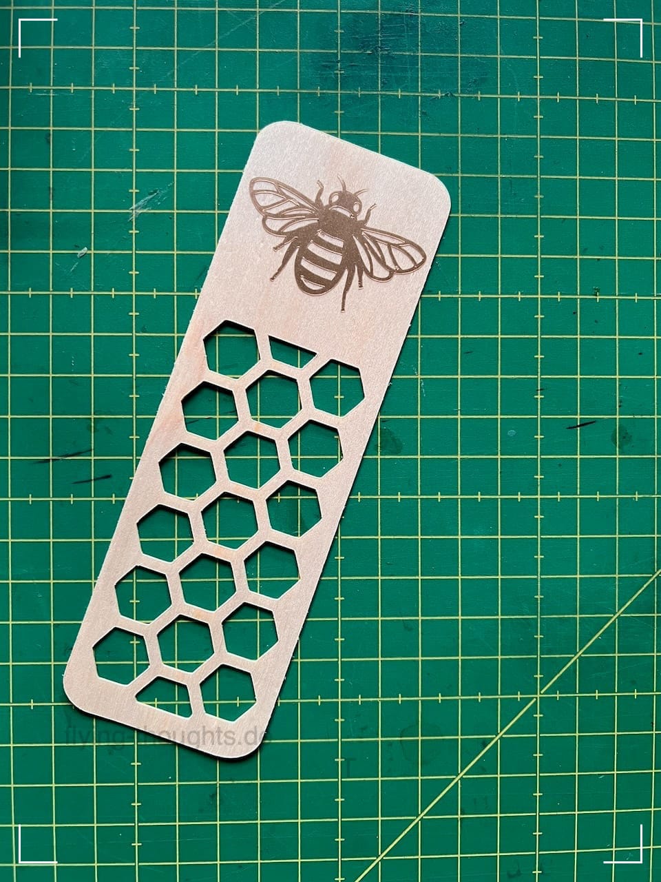 Ein Lesezeichen (Rechteck mit abgerundeten Ecken) aus Holzfurnier liegt auf einer grünen Schneidematte. 2/3 des Lesezeichens besteht aus ausgeschnittenen Sechsecken, die an Honigwaben erinnern. Im oberen Drittel ist eine Biene aus goldfarbenem Vinyl aufgeklebt. 