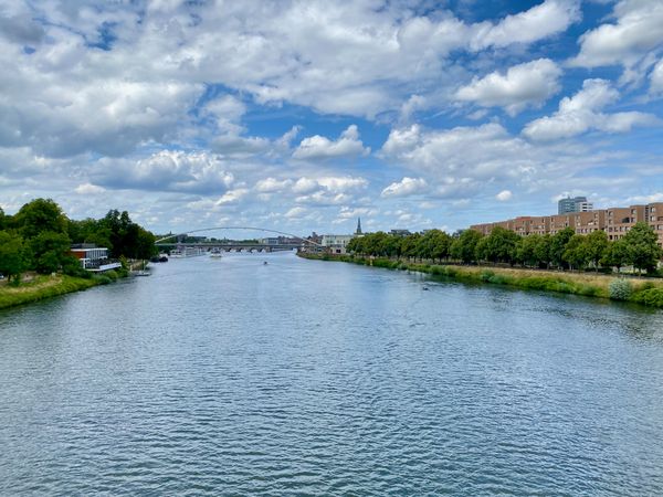 das Bild zeigt einen Fluss, aufgenommen von einer Brücke über dem Fluss. Links und rechts sind Bäume. Der Himmel ist bewölkt,