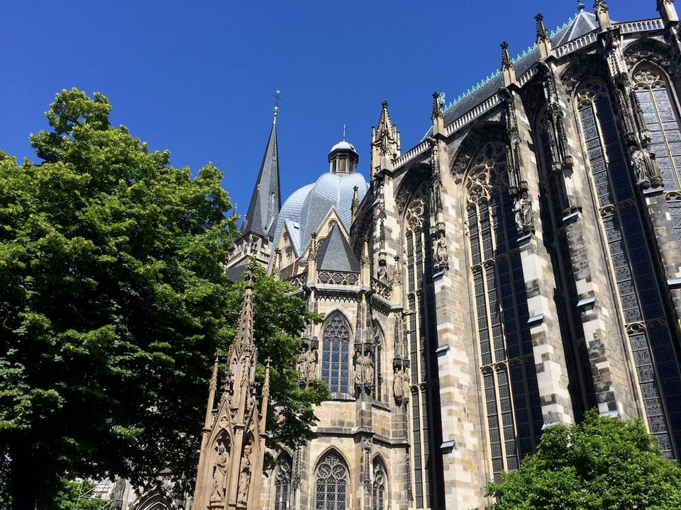 Grüne Bäume vor dem Aachener Dom. Der Himmel ist blau.